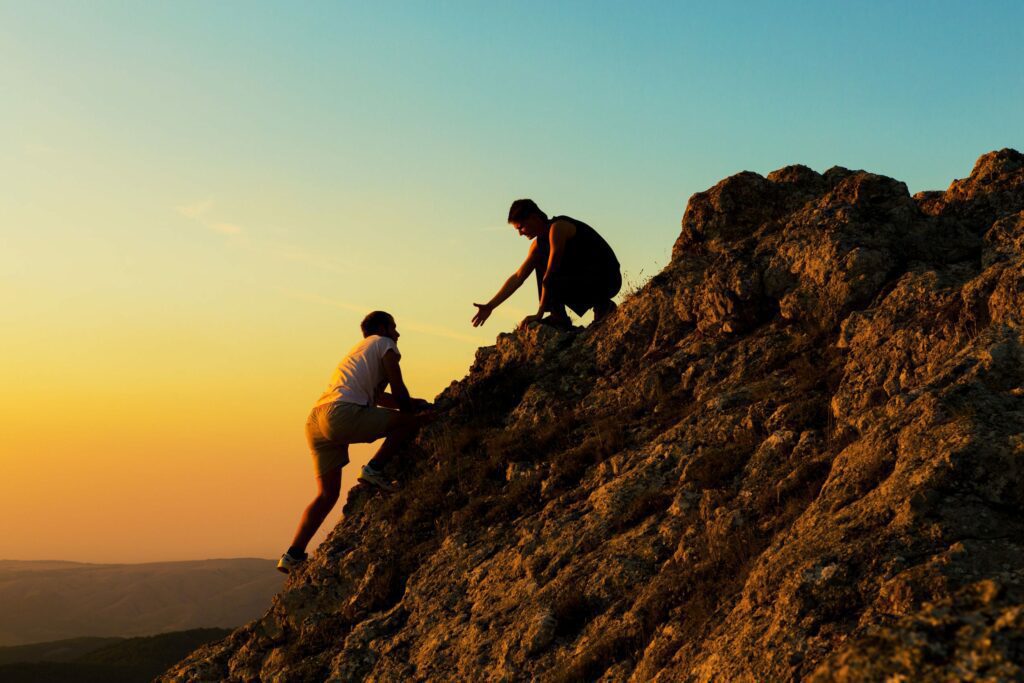 Two individuals are rock climbing at sunset. One climber extends a hand to assist the other on a rugged cliffside. The sky is gradient shades of orange and blue.