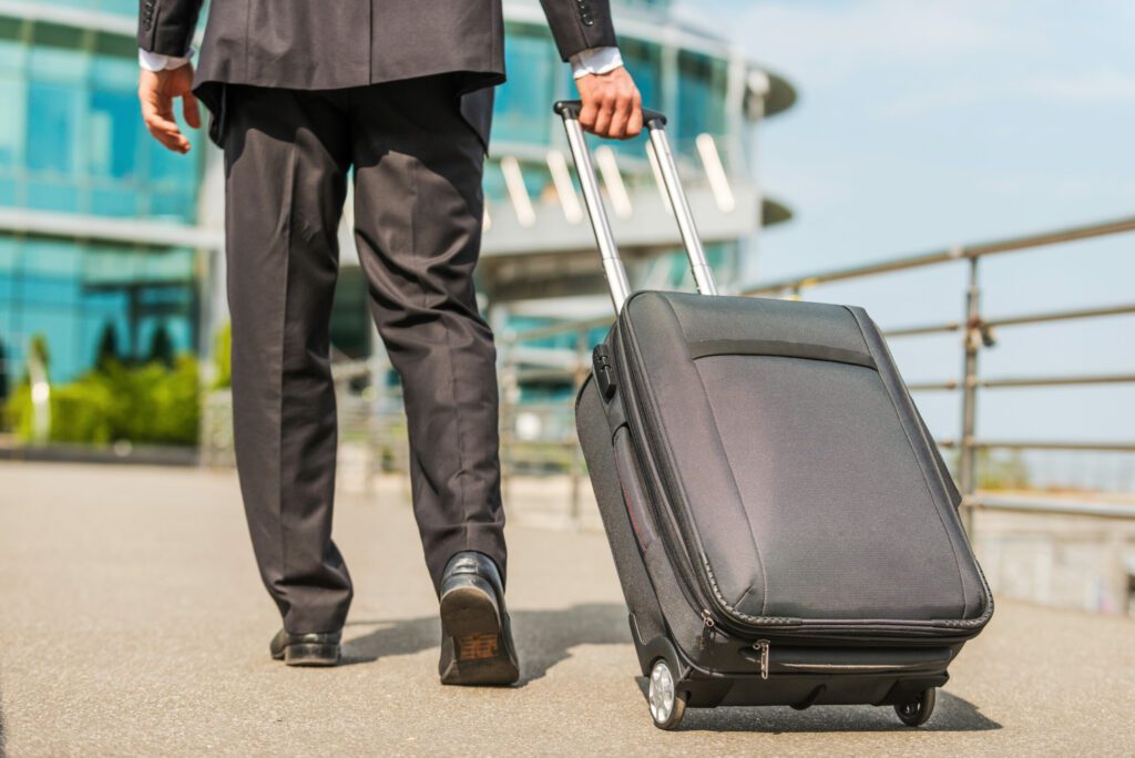 A businessman in a suit pulling a black wheeled suitcase outside a modern building.