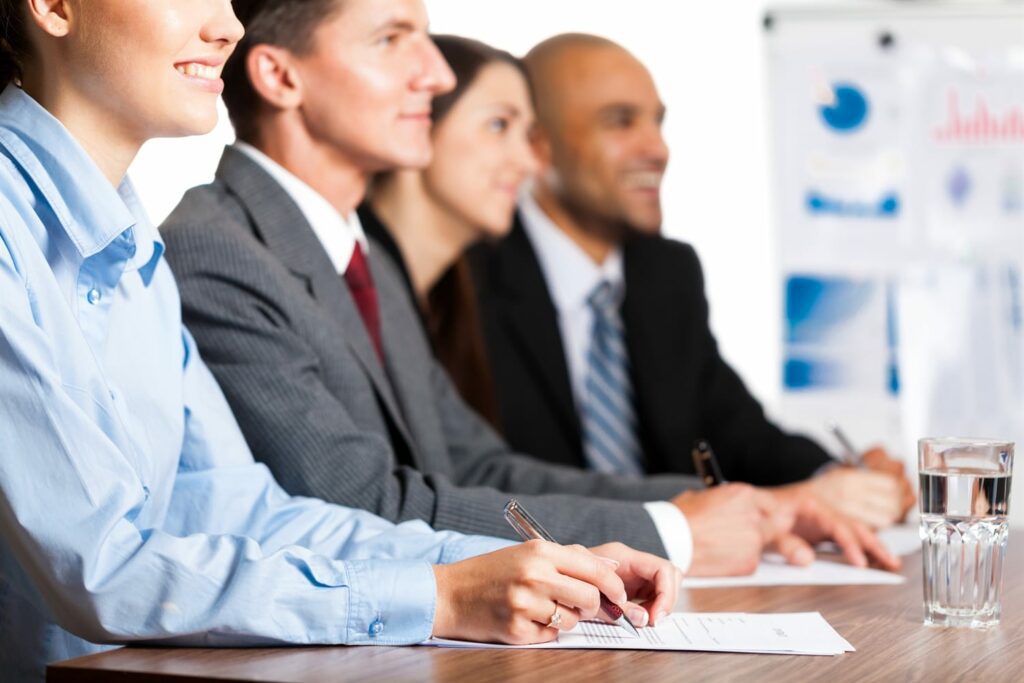 Group of five business professionals listening attentively at a conference table with water glasses and documents in front, and a chart board in the background.