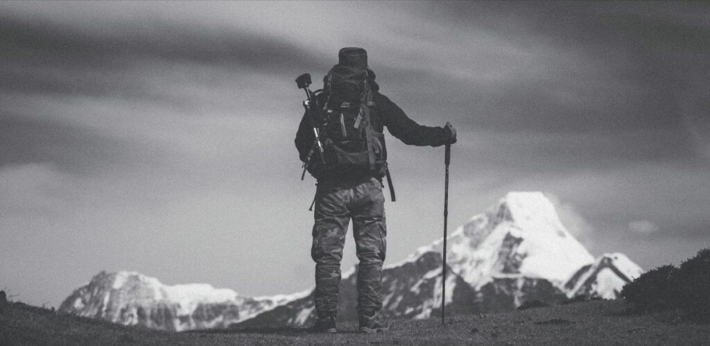 A hiker with a backpack and trekking pole stands facing a snow-capped mountain under a cloudy sky, ready to tackle the challenges ahead.