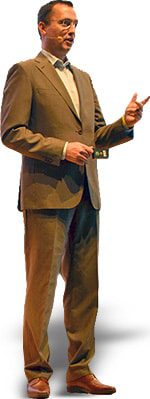 A man in a suit and tie gestures while speaking, standing confidently with one hand holding a pointer.