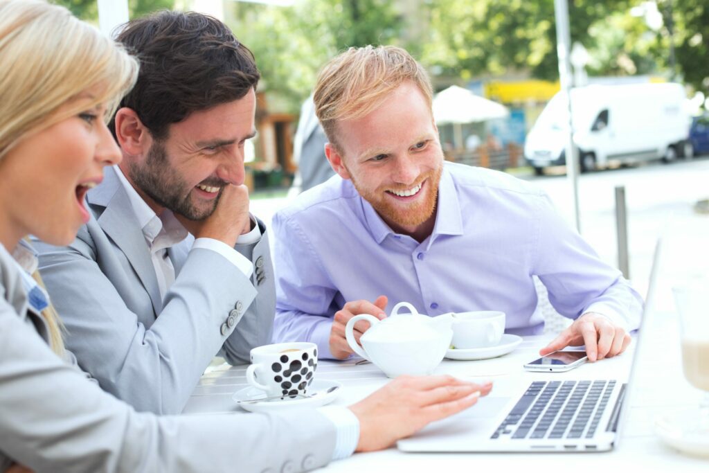 Three business colleagues, two men and one woman, laugh and look at a laptop at an outdoor cafe table with coffee cups.