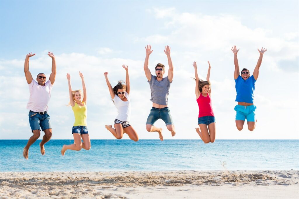 Six people jump in unison on a sandy beach with the ocean and a blue sky in the background. They are smiling and have their arms raised.
