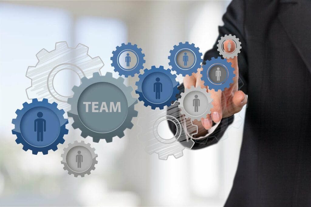 A person in a suit is touching interconnected gears, with icons of people inside each gear and the word "TEAM" in the central gear. The background is blurred and features light streaming in.