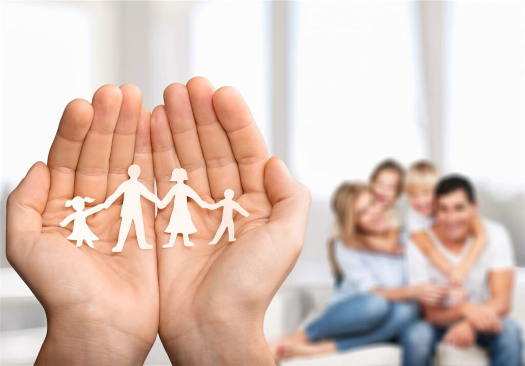 Close-up of hands holding paper cutouts of a family (two adults and two children) with an out-of-focus background of a smiling family of four sitting together.