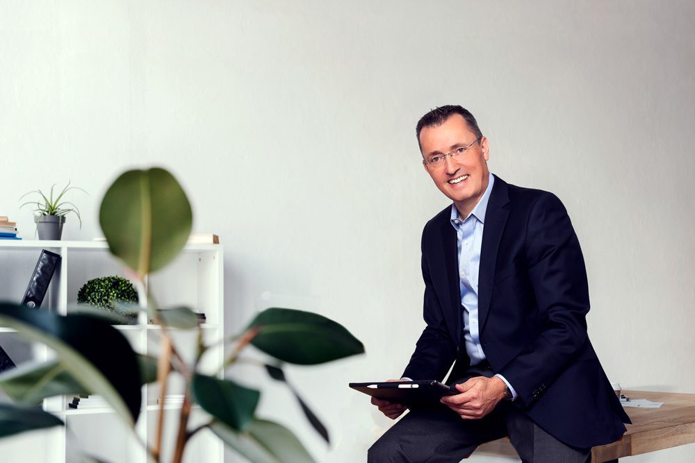Smiling middle-aged man in a suit sitting on a desk in an office environment holding a tablet.