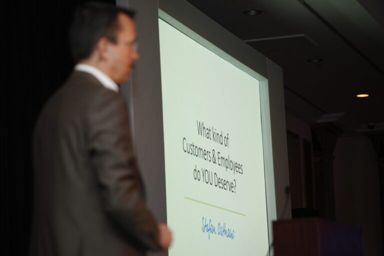 A person in a suit stands near a presentation screen displaying the question, "What kind of Customers & Employees do YOU Deserve?" with a signature below.