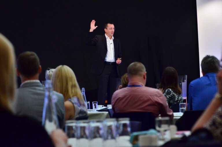 A man in a suit stands on a stage, raising one hand, addressing an audience seated at tables during a presentation or conference.