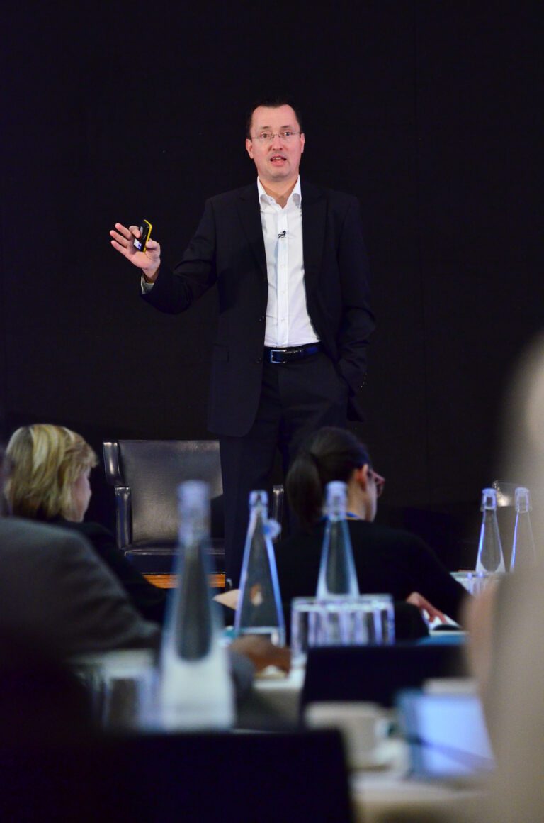 A man in a dark suit and white shirt stands on a stage, giving a presentation to an audience. He holds a clicker in one hand and gestures with the other.