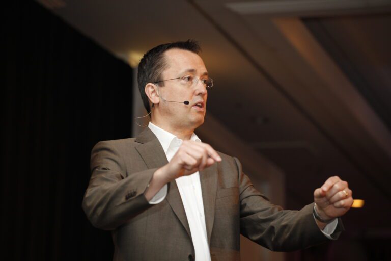 A man in a brown suit and white shirt speaks into a microphone headset while making hand gestures.