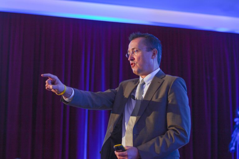 A man in a suit is speaking and pointing his finger during a presentation in a room with dark red curtains and blue overhead lighting.