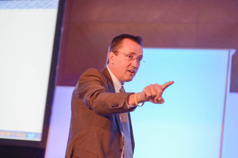 A man in a suit and tie gestures with his hand while speaking, standing in front of a blurred background with a screen.