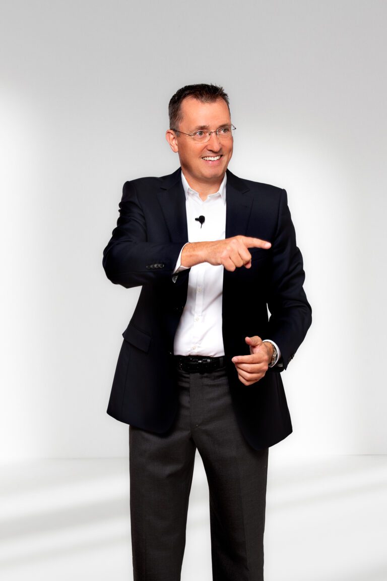 A man in a dark suit and white shirt is smiling and gesturing with his right hand while standing against a plain white background.