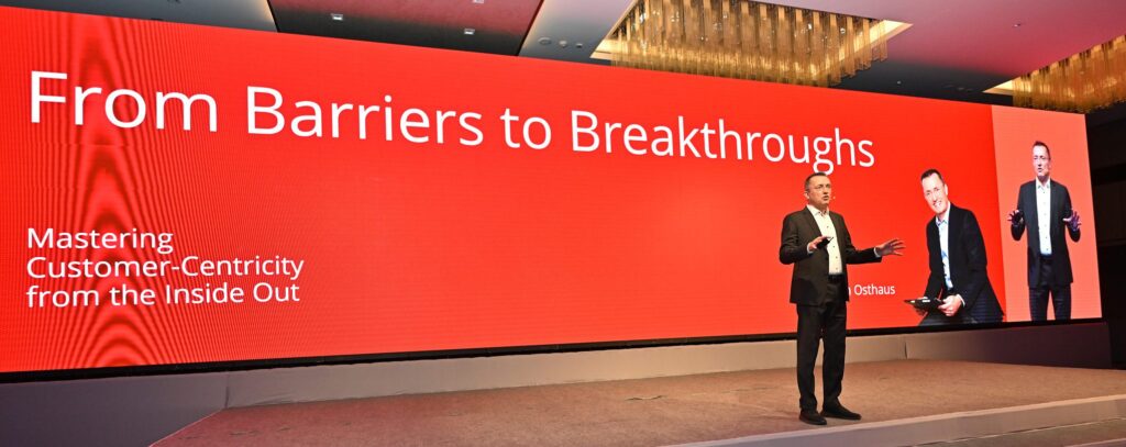 A man stands on stage in front of a large red screen displaying the text, "From Barriers to Breakthroughs: Mastering Customer-Centricity from the Inside Out.