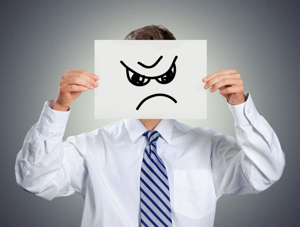 A man in a white shirt and blue tie holds a paper with a drawn angry face covering his own.