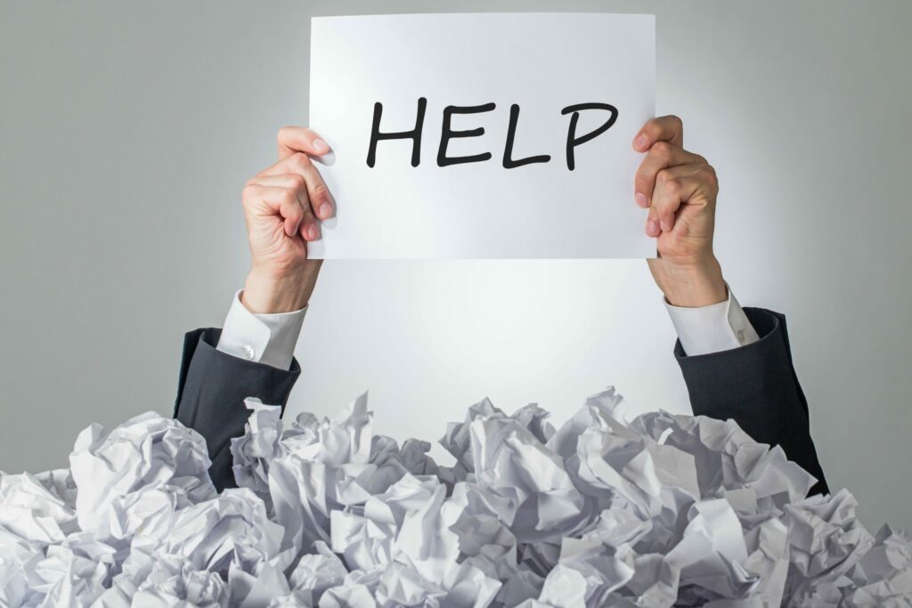 A person submerged in crumpled paper holds a sign reading "help" above their head.