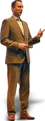 A man in a suit and tie gestures while speaking, standing confidently with one hand holding a pointer.