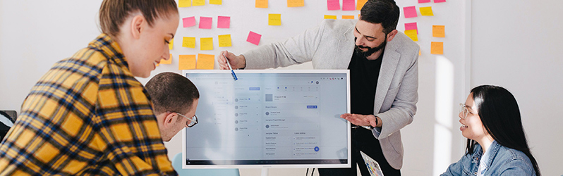 Four professionals collaborating around a digital screen, discussing a project in a meeting room with sticky notes on the wall.