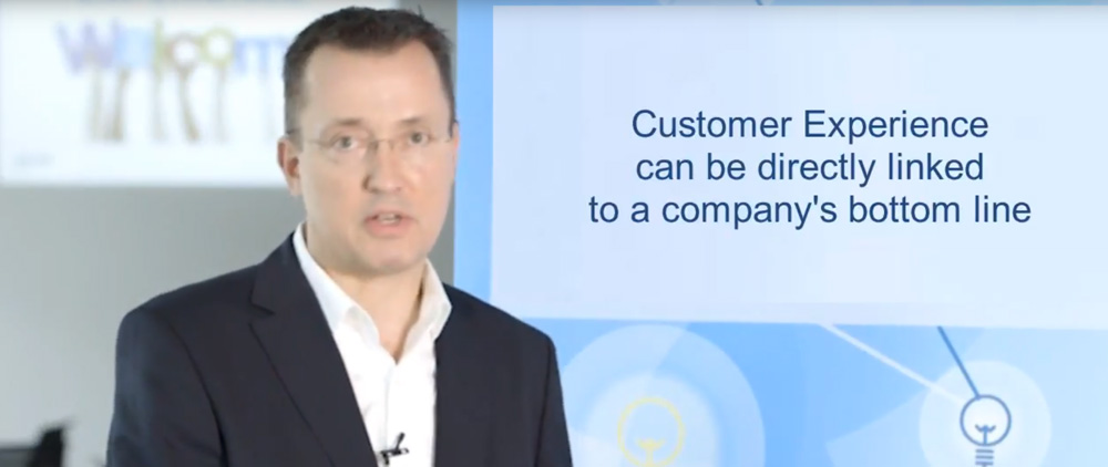 A man in a white shirt presenting a slide about "customer experience" relating to a company's bottom line in a conference room.