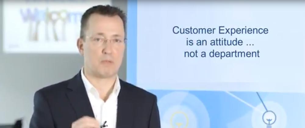 A man in a suit and glasses speaking at a seminar with a presentation slide in the background reading "customer experience is an attitude ... not a department.