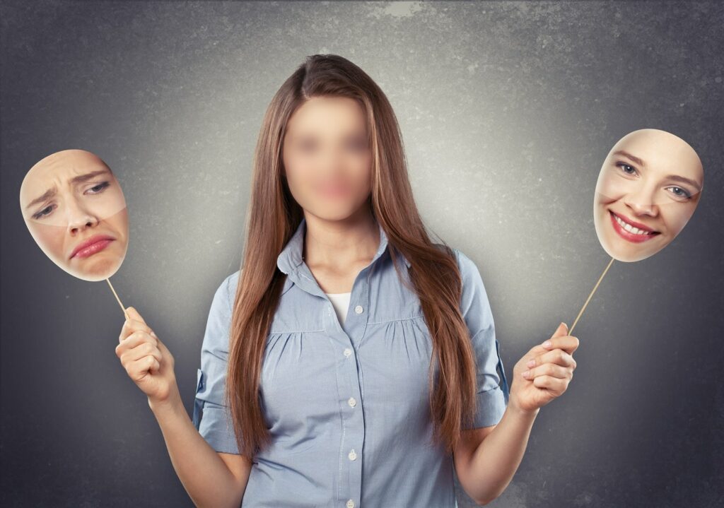 A woman with a blurred face is holding two masks on sticks; one mask shows a sad expression, and the other mask shows a happy expression.