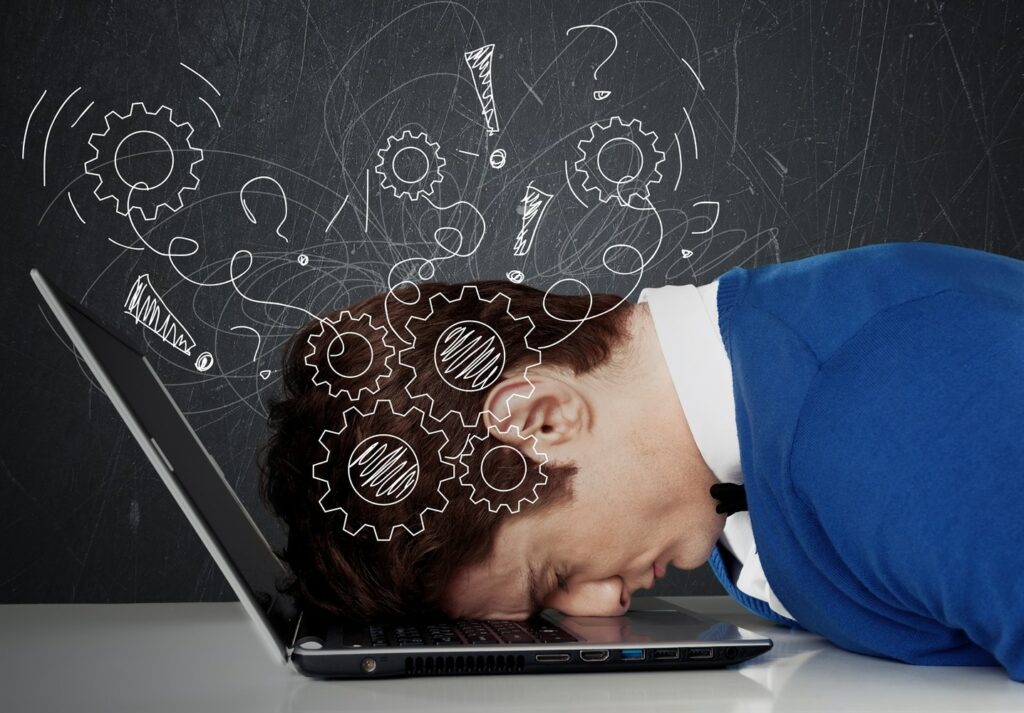 Person in a blue sweater with their head down on a laptop keyboard, surrounded by scribbled gears and question marks on a blackboard in the background.